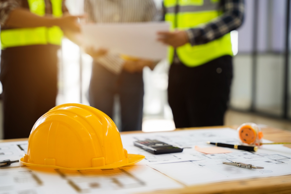 Yellow,Hard,Hat,On,Workbench,With,Colleagues,Discussing,Data,Working