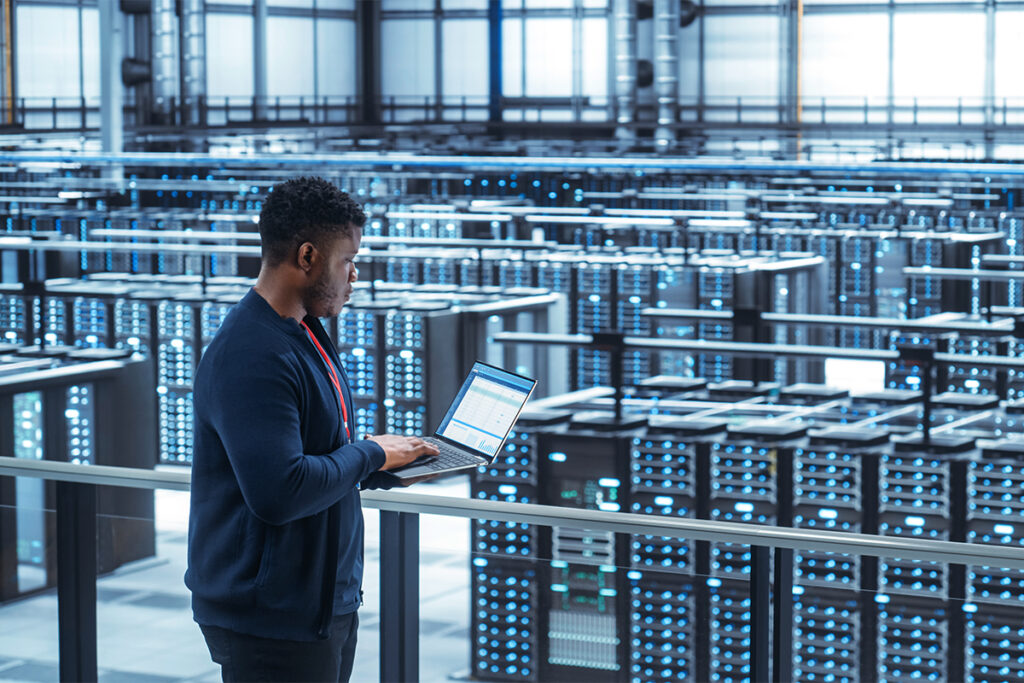 Data Center Engineer Using Laptop Computer. Server Farm Cloud Computing Specialist Facility with African American Male System Administrator Working with Data Protection Network for Cyber Security.