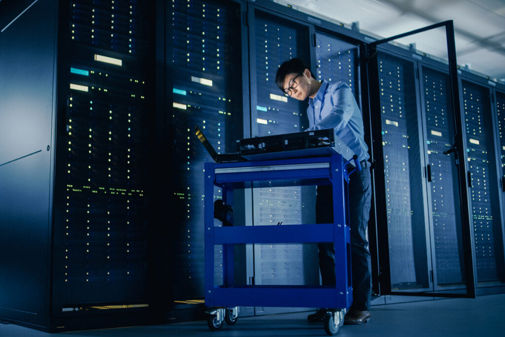 In Data Center: Male IT Technician Running Maintenance Programme on a Laptop, Controls Operational Server Rack Optimal Functioning. Modern High Tech Telecommunications Operational Super Computer.